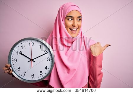 Young beautiful arab woman wearing pink muslim hijab doing countdown holding big clock pointing and showing with thumb up to the side with happy face smiling