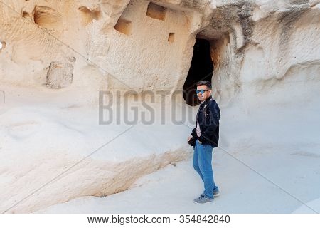 Man In A Black Jacket And Jeans, Wearing Sunglasses, Stands At The Cave. Cappadocia Turkey.