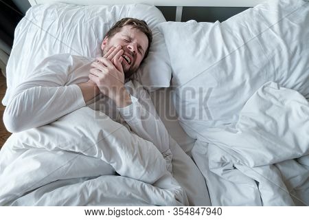 Man In White Pajamas Suffers From Severe Toothache, He Holds Hand On His Cheek, Lying In Bed, In The