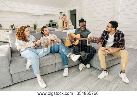 Young Africam man playing guitar while sitting on sofa among his friends listening to music while relaxing in living-room