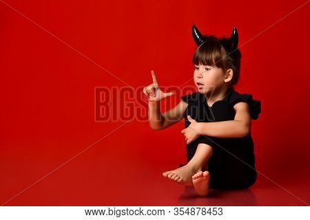 Little Brunette Girl With Devil Horns, In Black Blouse And Leggings, Barefoot. She Is Looking At Her