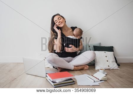 Young Freelance Mother Having Her Baby In Ergo Backpack And Working At Home Using Laptop And Smart P