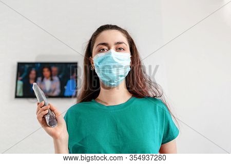 A Woman In Medical Mask Holding A Remote Control From The Tv. In The Background A White Wall With Tv