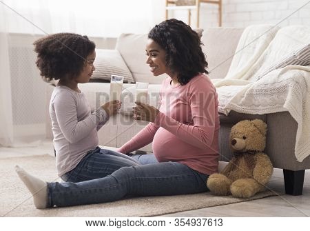 Healthy Drink. Little Afro Girl And Her Pregnant Mother Drinking Milk Sitting On Floor At Home, Bond