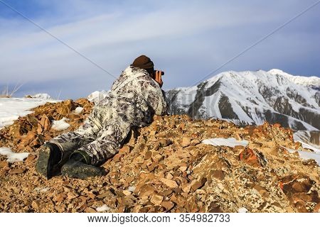 Hunter In Winter Camouflage In The Mountains Lying Watching With Binoculars. A Huntsman Lies In The 