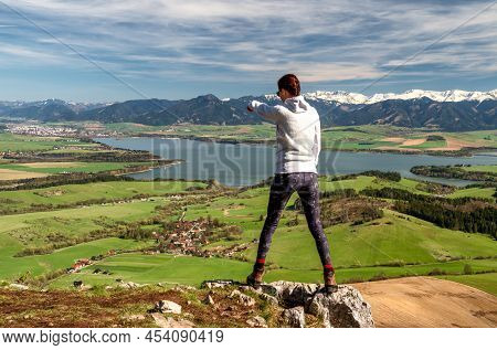 Woman Hiker Standing On Top Of The Hill And Looking On Nature With Lake And Snowy Mountains In Regio