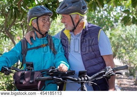 Active Senior Caucasian Couple With Electrobikes Standing Outdoors In The Park. Two Happy Elderly Pe