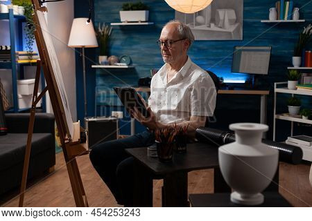 Senior Man Art Creator Browsing Digital Tablet Looking For Inspiration In Online Art Gallery In Home