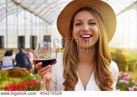Close-up Of Young Woman Looking At Camera And Holding Glass Of Wine With People Among Flowers Field 