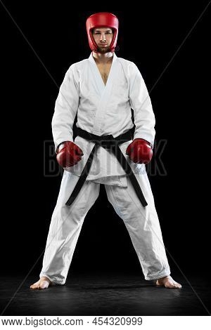 Portrait Of Male Taekwondo Fighter In White Dobok, Helmet And Gloves Posing Isolated Over Dark Backg
