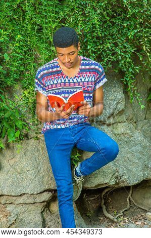 Man Reading Outside. Wearing A Short Sleeve, Collarless, Colorful Pattern Shirt, Blue Jeans, A Young