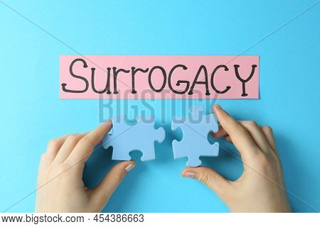 Woman Holding Puzzle Pieces Near Sheet Of Paper With Word Surrogacy On Light Blue Background, Top Vi