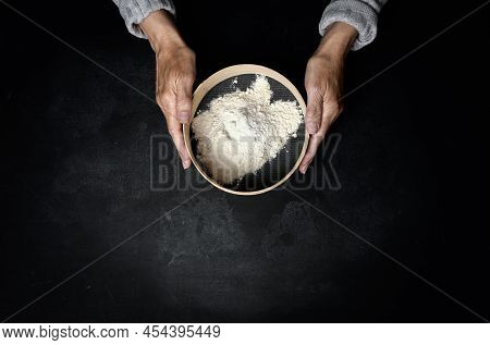 Two Female Hands Hold A Round Wooden Sieve With Wheat Flour On A Black Table, Top View