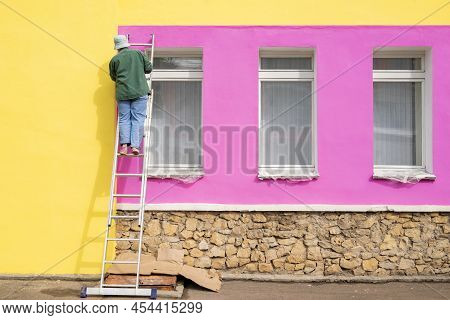 Female Painter Paints The Wall Of The Facade Of A Building While Standing On The Stairs. The Concept