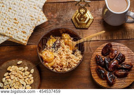 A Plate Of Couscous With Fruits And Nuts On A Wooden Table Next To Matzah And A Jug Of Fruit Broth. 