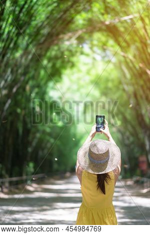 Asian Woman In Yellow Dress And Hat Traveling At Green Bamboo Tunnel, Happy Traveler Taking Photo By