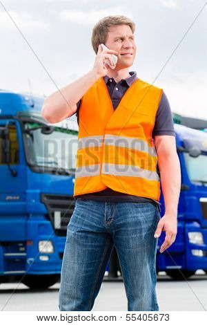 Logistics - proud driver or forwarder with mobile phone in front of trucks and trailers, on a transshipment point