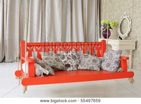 Big red wooden swing with pillows and  console mirror in stylish light studio.