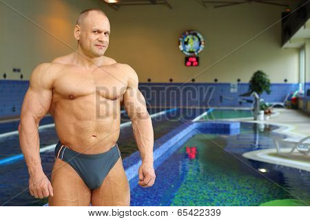 Bodybuilder in swimming trunks stands near indoor pool of gym hall