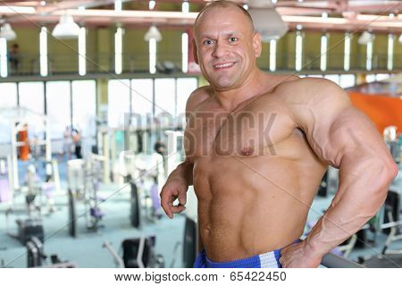 Happy shirtless sunburnt bodybuilder stands in big modern gym hall