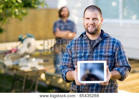 Portrait of mid adult carpenter displaying digital tablet with coworker in background at construction site