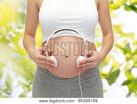 pregnancy, people, music, technology and expectation concept - close up of happy pregnant woman applying headphones to bare tummy over green tree leavers background