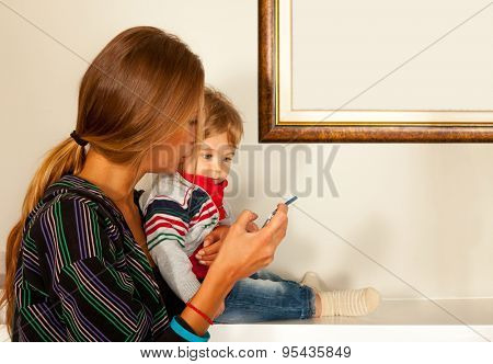Young beautiful mother and a cute blue eyes child are looking at a smartfone indoors. Copy space frame.