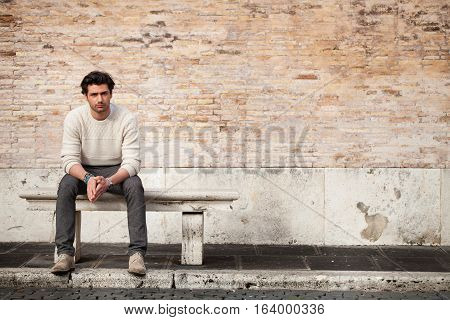 Handsome young man sitting on marble bench with bricks background. A beautiful and young boy sat on a marble bench. Behind him a large, old brick wall. Beside him, a large copyspace for custom text or graphic