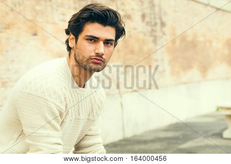 Handsome beautiful young man outdoor. Fashion hairstyle. Handsome young boy looking. Stylish hair, charming and attractive look. Slight beard and white sweater. Outdoors in the city. Clear and bright environment.