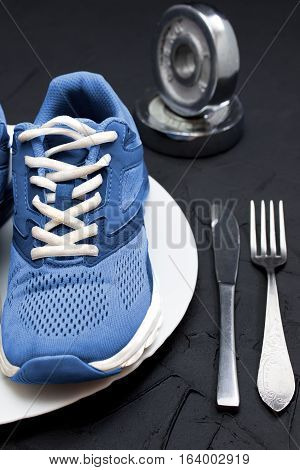Blue sport shoes on wtite plate fork and knife on black concrete background vertical orientation. Concept healthy lifestyle sport and diet. Focus is only on the sport shoes.