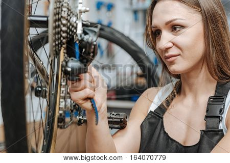 Delighted from my job. Cheerful smiling skillful craftswoman standing in the repair shop and working while fixing the chain of the bicycle