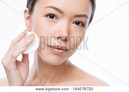Pretty cheerful asian woman pressing a cotton pad to her cheek and cleaning her face while caring about skin