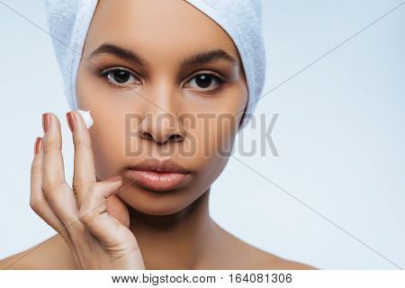 For shiny skin. Attractive pretty nice woman having some moisturizing cream on her finger and preparing to apply it while wearing towel turban