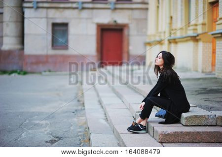 Young attractive long-haired girl student in black jacket jeans and sneakers thoughtfully sitting on stairs in yard.