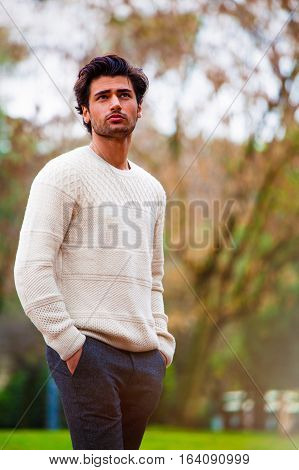 Fashion man. Beautiful young male. Handsome boy outdoors. Beautiful man outdoors. The young man is in a park and is dressing a white sweater and gray trousers. Exciting look and hands in pockets. Italian.