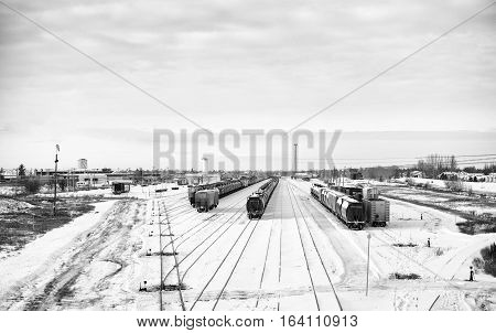 Industrial railway shipping cars docked at a yard site surrounded by houses and industrial buildings on the outskirts of a city in black and white winter landscape