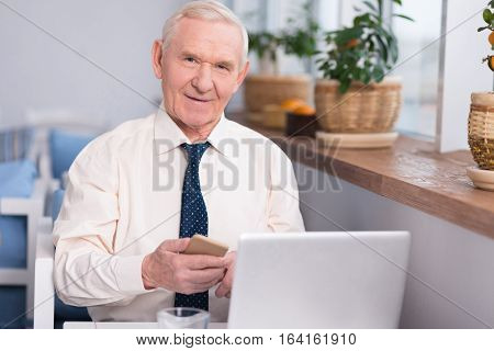 I will inform you. Pleasant cute mature businessman calling someone on his phone while sitting at the table in front of his computer