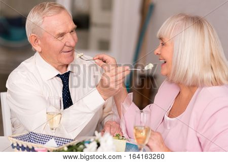 Precious moments. Lovely radiant senior couple giving each other bites of cake while having fun celebrating their anniversary in cafe