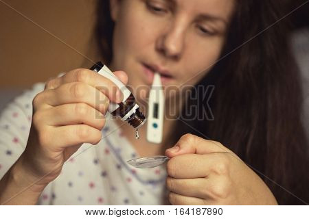 Woman with a thermometer in his mouth dripping curative medicine in a spoon