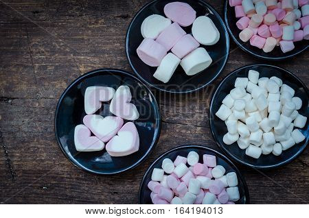Colorful mini marshmallows in a black plates on a wooden background. Different mini white pink and orange puffy marshmallows. Marshmallow concept.