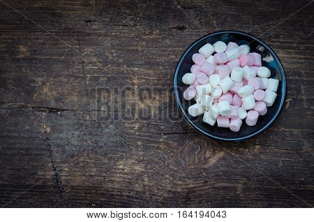 Pink and white mini marshmallows in a black plate on a wooden background with place for text. Different mini puffy marshmallows. Marshmallow concept. Top view. Copy space.