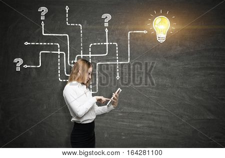 Side view of a girl with a tablet working near a blackboard with an arrow maze and a light bulb sketch on it. Concept of finding the answer