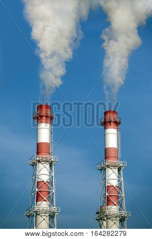 Two striped red and white industrial pipes with white smoke over cloudless blue sky. Front view closeup