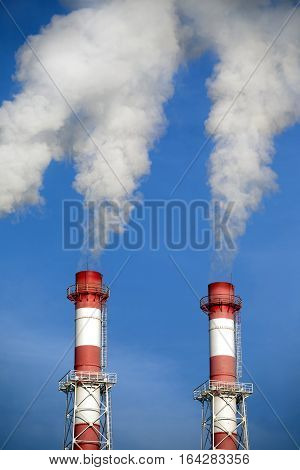 Two striped red and white industrial pipes with white smoke over cloudless blue sky. Front view closeup