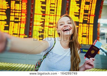 Girl In International Airport, Taking Funny Selfie With Passport And Boarding Pass Near Flight Infor