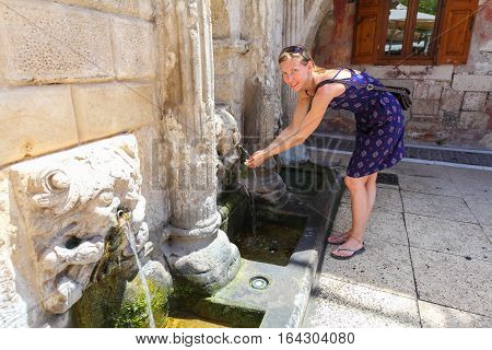 Rethymnon, Island Crete, Greece - July 1 2016: Young woman is drinking clear cold water from Rimondi Fountain in Rethymnon (part of Old Town) in hot summer day. The Rimondi fountain was built in 1626 by the city's Rector A. Rimondi.