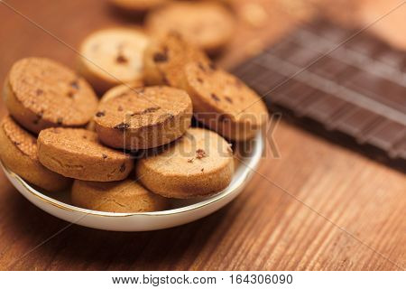 Beautiful tasty biscuit cookies with chocolate chips standing in plate on wooden table with crumbs and chocolate bar at the background. Shallow dof. Focus on foreground.