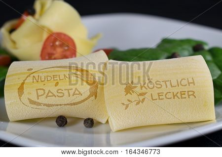 On a white plate are 2 noodles with the lettering "Noodles Patsa - Exquisite Delicious" in german language in the foreground. In the background are further noodles garnished with leaf spinach tomatoes and pepper grains