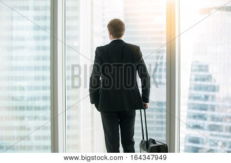 Young businessman with suitcase standing looking at the full length window, before a trip undertaken for work or business purposes, leader with high business aspirations. Rear view