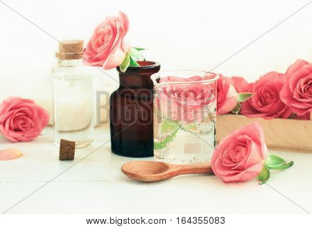 Rose cosmetic ingredients for herbal spa beauty treatment. Flower in attar glass test jar with ml scale on wooden white table. Soft focus, soft light.